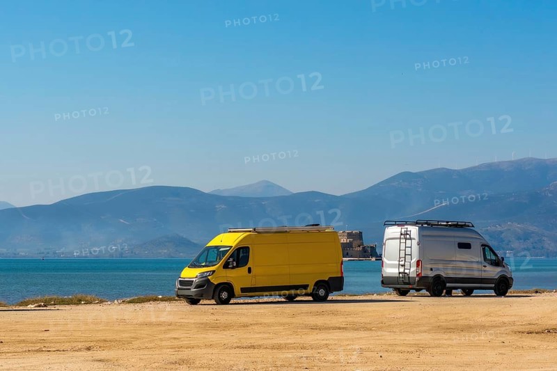 Two camper vans parked at Arvanitias Beach in Nafplion, Greece, with the Palamidi Fortress on the mountain in the background. A perfect spot for van life and coastal exploration