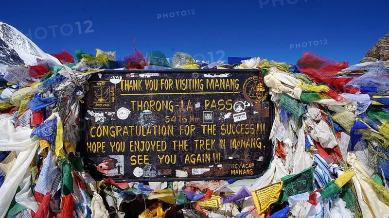 Prayer flags and sign on Thorong La Pass in Manang in front of snow-capped mountains, trekking at Annapurna Circuit, Nepal