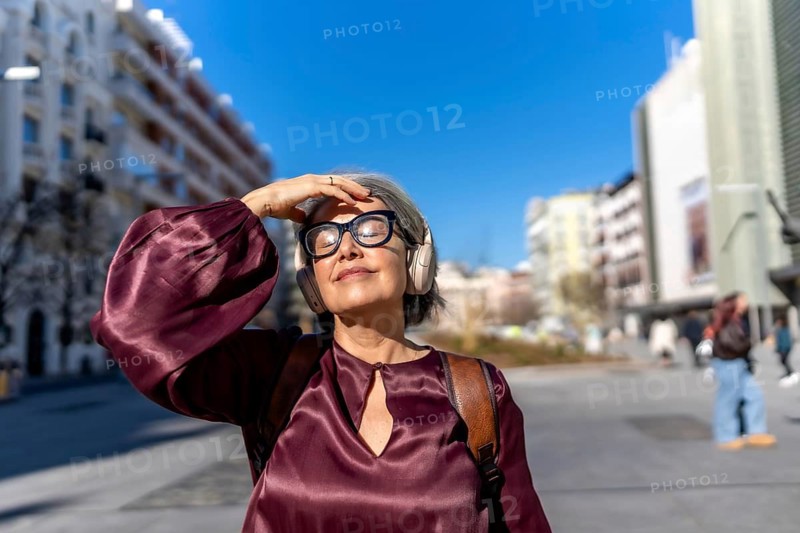 Mature woman with gray hair wearing headphones and eyeglasses, shielding her eyes from the bright sun while relaxing and listening to music outdoors in a bustling urban environment