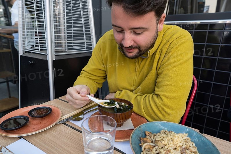 A man in a yellow sweater enjoys a traditional Asian meal at an outdoor restaurant table. He uses a spoon to taste soup, surrounded by dishes and drinks