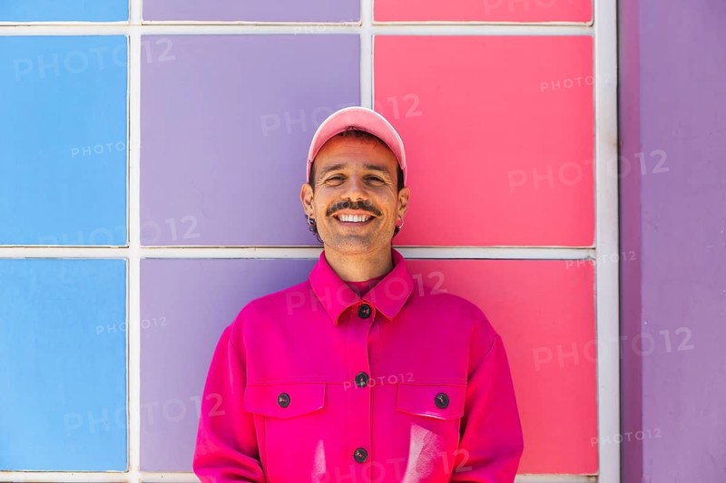A young Spanish man with a mustache, wearing vibrant pink clothing, smiles joyfully against a colorful tiled background in Quito, Ecuador. The essence of urban lifestyle and fashion, alongside themes of self-expression with the LGTBIQ+ community