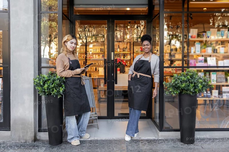 Two employees in aprons stand smiling at the entrance of a chic cosmetics store. The store's interior with products can be seen through the glass. A welcoming retail environment