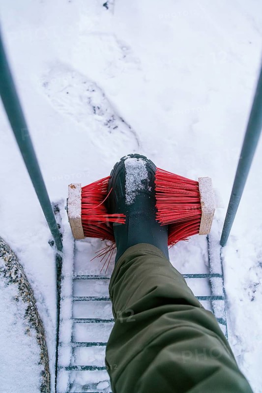 A person cleans snow off their boot using a red brush, set against a snowy backdrop in Swedish Lapland. The practical winter tool helps maintain cleanliness