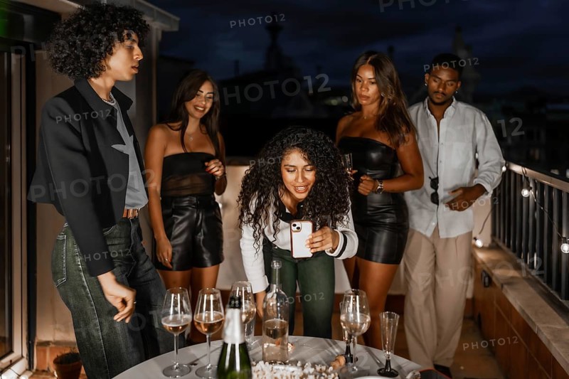 Woman using mobile phone during night party with multi-ethnic friends in a private rooftop with city views at night