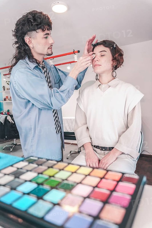 Professional makeup artist carefully applying eyeshadow on model's face using a brush, with a colorful palette of eyeshadows in foreground, creating a diverse and inclusive beauty experience