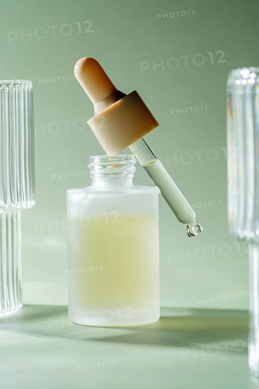 A dropper releasing a single drop of a translucent serum into a matte glass bottle on a serene green backdrop, symbolizing skincare and beauty