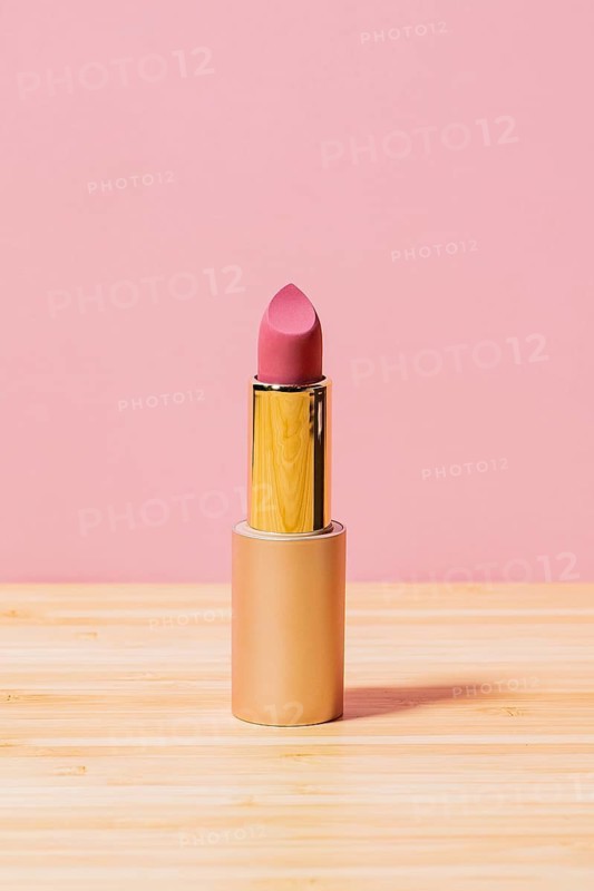 A single pink lipstick in an elegant golden tube placed on a wooden tabletop, set against a soft pink background, highlighting the product's sophisticated design
