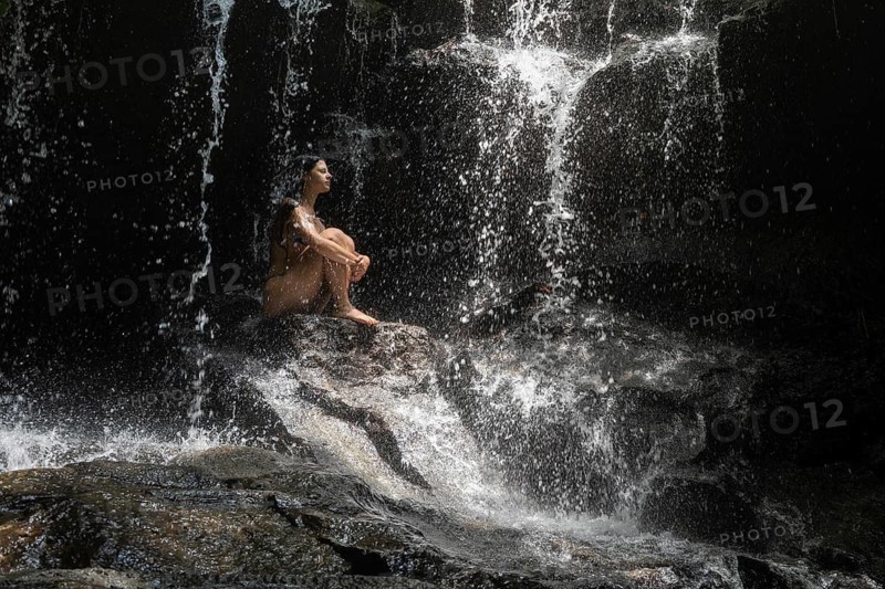 A serene scene of a woman sitting alone on rocks under a gentle waterfall, enveloped by nature of Bali. The tranquil moment emphasizes solitude, calmness, and reflection