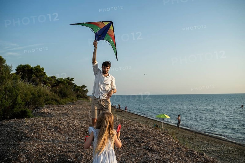 A father and daughter enjoy a playful moment on a scenic Italian beach. The father lifts a colorful kite into the sky while his daughter watches eagerly, creating a joyful summer memory