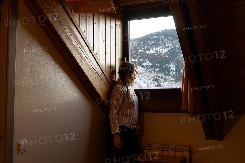 A woman with red hair looks away from the camera, standing by a window in a wooden attic, gazing at the snowy mountain landscape outside