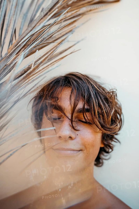 A relaxed teenage boy with tousled hair closes his eyes peacefully, partially obscured by dried palm leaves against a light background, creating a serene and calming scene