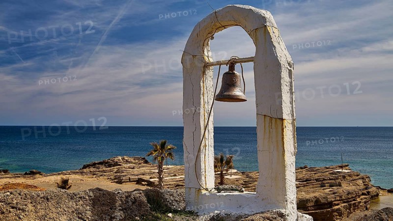 A rusty bell hangs under a white arch by the sea in front of a clear sky and rocks, Chapel, Agios Nikolaos, Rock Chapel, Oasis Beach, East Coast Rhodes, Rhodes, Dodecanese, Greek Islands, Greece