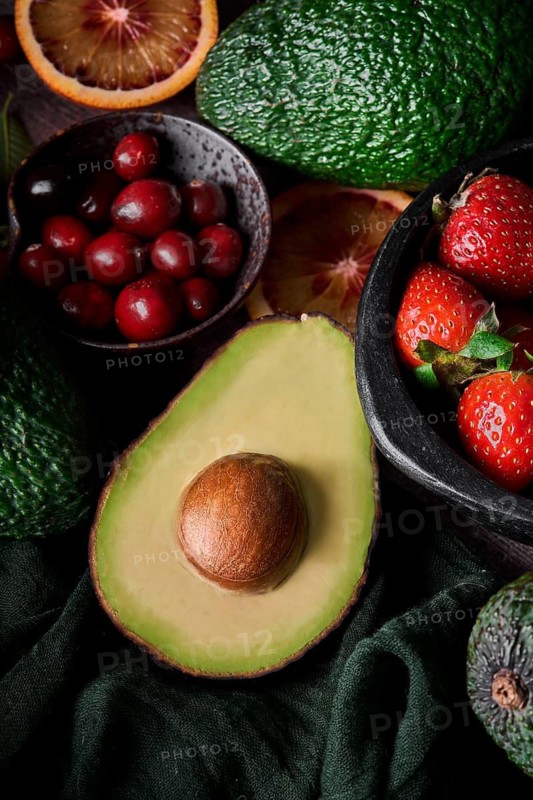 A fresh assortment of fruits including avocado, strawberries, cranberries, and orange slices displayed on a dark background, showcasing a vibrant and healthy selection