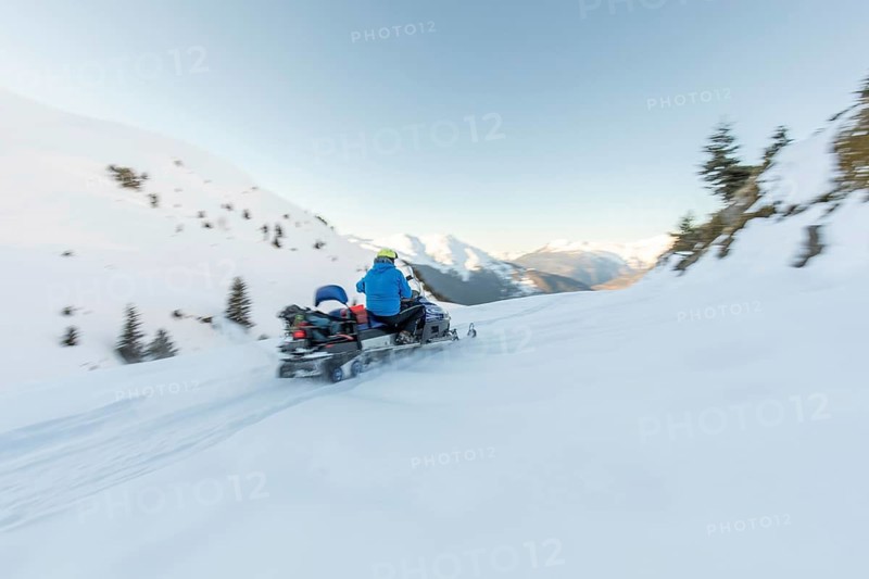 A snowmobiler in a blue jacket speeds through pristine snow-covered mountains, capturing the thrill of winter adventure against a backdrop of clear blue skies and rugged peaks
