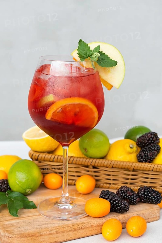 A glass of red sangria filled with citrus slices and mint sits on a wooden board. Surrounding the drink, an assortment of blackberries and citrus fruits in a wicker basket enhance the summer vibe