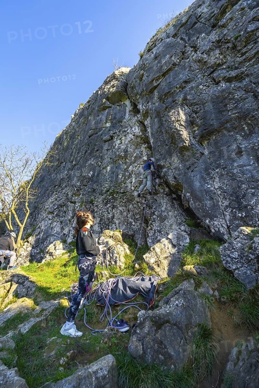 Two female rock climbers ascending a challenging rock face in nature, one belaying from below while the other climbs higher