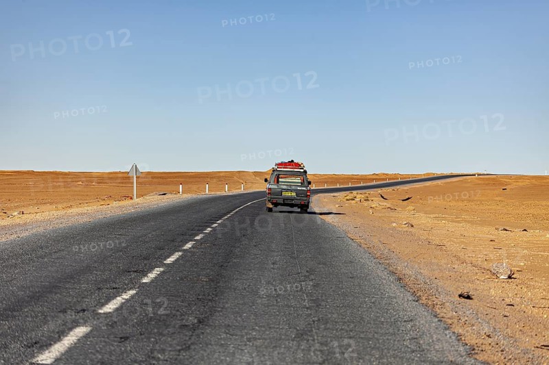 Lonely road with a vehicle in the distance, surrounded by empty sandy areas, under a clear sky, By car through the Sahara desert in Algeria