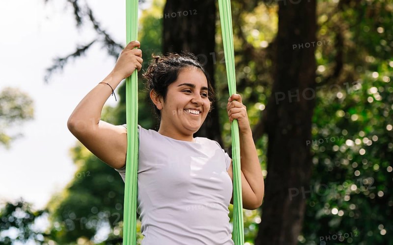 A latin woman joyfully practices aerial acrobatics on fabric in a lush Bogota park, surrounded by nature Their expression reflects excitement and focus in this dynamic outdoor setting