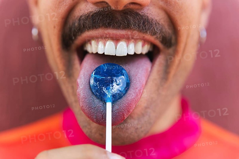 Cropped unrecognizable LTBTIQ+ man with a mustache is depicted enjoying a blue lollipop in Quito, Ecuador. Dressed in vibrant pink, he embodies a cheerful, urban lifestyle and represents diversity and self-expression