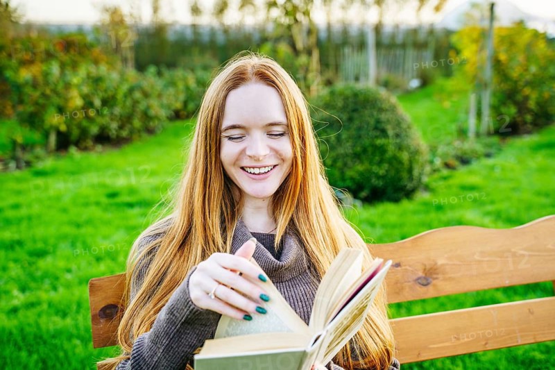 A beautiful pregnant woman with red hair enjoys reading a book on a wooden bench in a lush green park, basking in the warm sunlight and embodying serenity and joy
