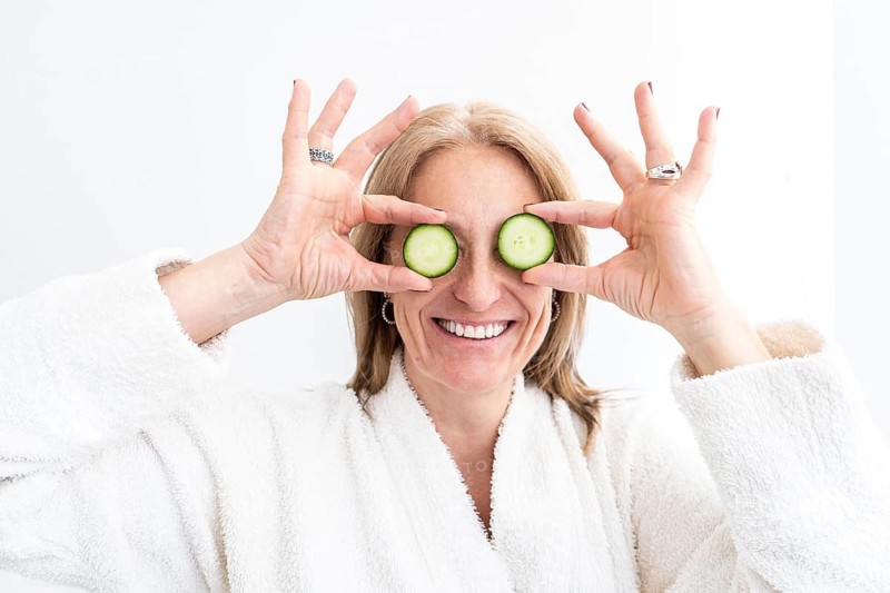 Mid-adult woman in a spa setting holding cucumber slices over her eyes, wearing a white robe. The bright background adds to the serene atmosphere