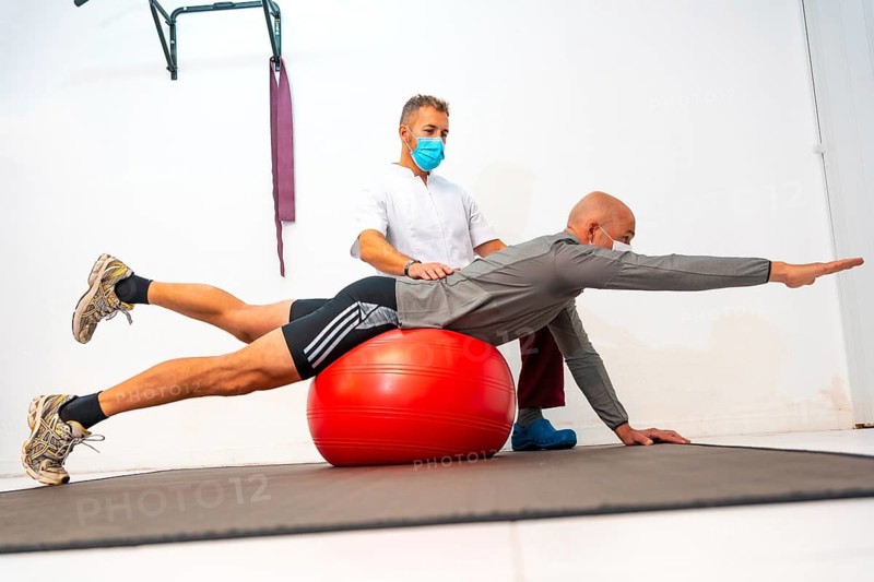 Physiotherapist with face mask helping client to balance on a red ball. Physiotherapy with protective measures for the Coronavirus pandemic, COVID-19. Osteopathy, sports quiromassage
