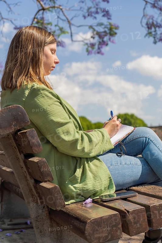 A vertical shot of a young woman sitting on a bench in a park, writing in a notebook with a pen. The scene includes vibrant trees, flowers, and a sunny sky, evoking creativity and relaxation