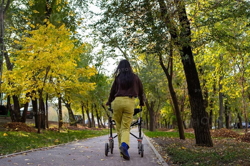 A woman with cerebral palsy walks with a walker on a scenic park path surrounded by vibrant autumn trees. The colorful foliage creates a serene and peaceful atmosphere