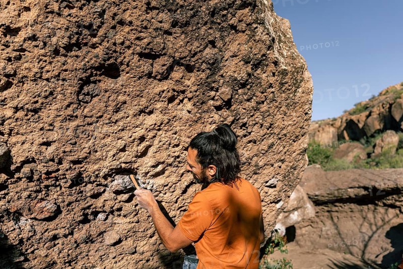 A climber tackles a rugged boulder, demonstrating skill and focus in a natural setting. The sunlit scene highlights the rocky texture and untouched wilderness around