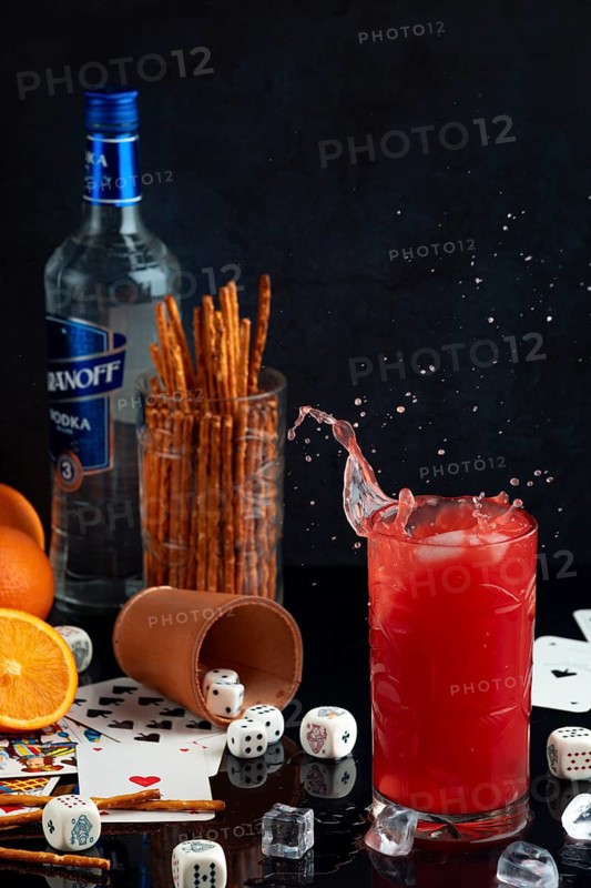 Splashing long drink with vodka and oranges, surrounded by cards, dice and pretzel sticks, studio shot