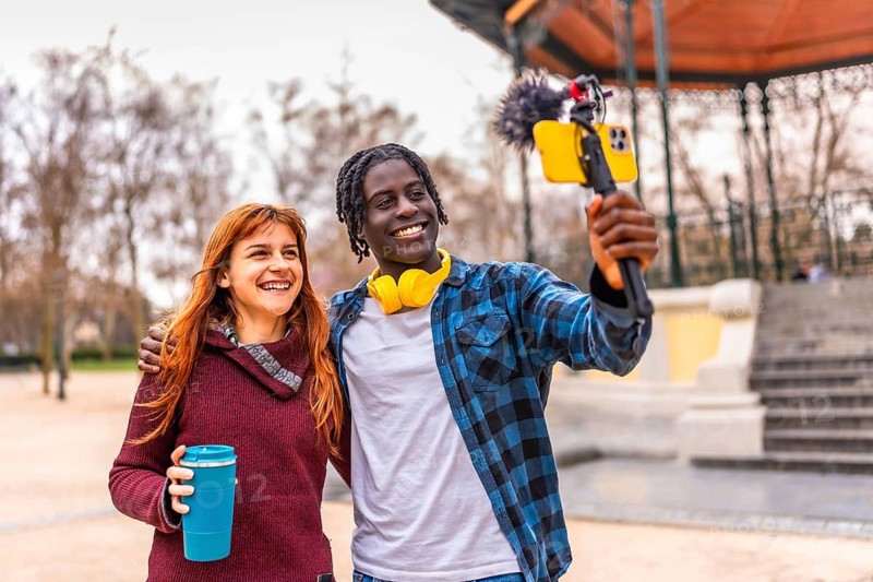 Two young content creators are recording a video blog using a smartphone with a microphone in a park
