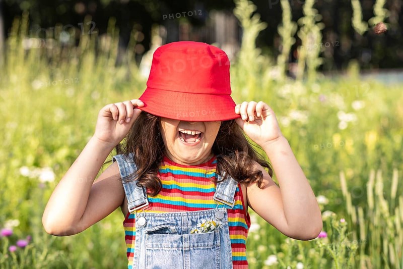 A cheerful little girl playfully hides behind a bright red hat, laughing joyfully in a sunlit meadow filled with vibrant flowers, embodying happiness and childhood innocence
