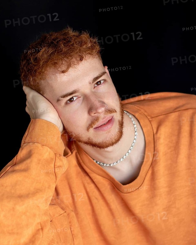 A young man with red hair and a gentle expression, wearing an orange shirt and pearl necklace. He poses thoughtfully against a dark backdrop, hand resting on his face