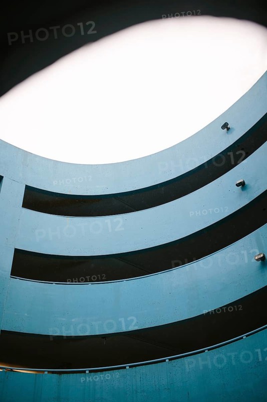 A modern, blue parking lot featuring a multi level circular design enhanced by soft lighting from an overhead aperture. The architectural detail focuses on the curved lines and shadows