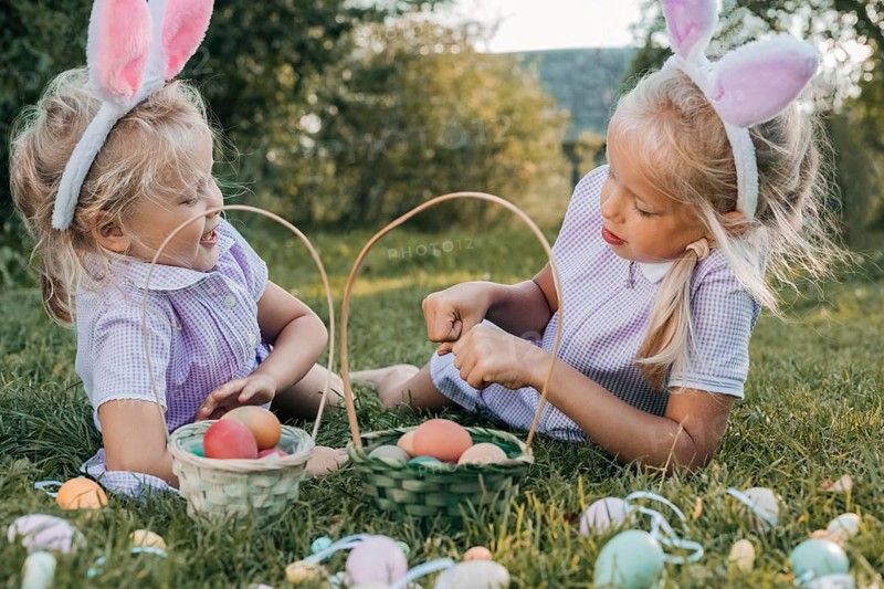 Two cheerful children with bunny ears sit on grass, baskets full of colorful eggs, enjoying a playful Easter egg hunt in a sunny meadow surrounded by greenery