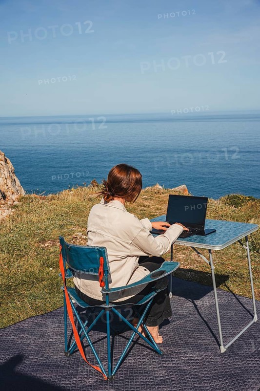 A woman enjoys remote work while sitting on a chair near the ocean, surrounded by stunning views. An ideal setup for digital nomads traveling in a camper