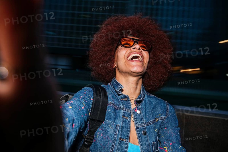 A joyful African descent woman with afro hair, wearing orange sunglasses, laughs heartily in an urban environment. She is casually dressed in a denim jacket and carries a backpack