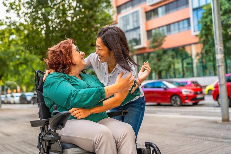 Woman with disability in wheelchair and multi-ethnic friend reunited embracing in the city street