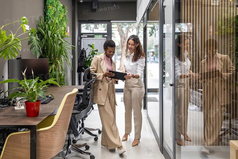 Two colleagues in a modern office engage in a discussion, using a digital tablet. Surrounded by plants and glass partitions, reflecting a collaborative and eco-friendly workspace