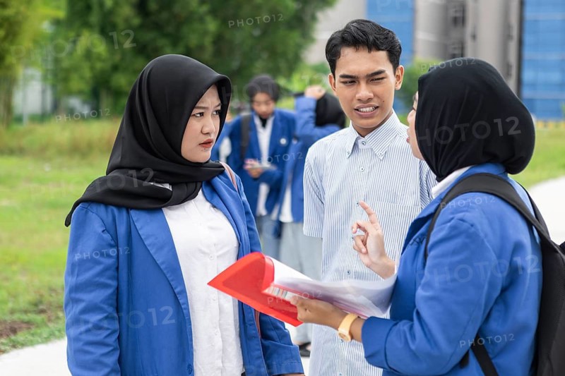 A group of Asian Muslim students in blue uniforms engaging in a conversation outdoors. They stand on a path surrounded by greenery, indicating a school environment
