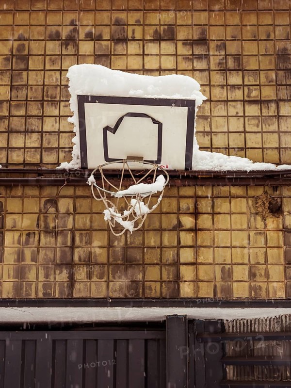 A basketball hoop laden with snow, backed by a grid of brown tiles, evoking urban wintertime