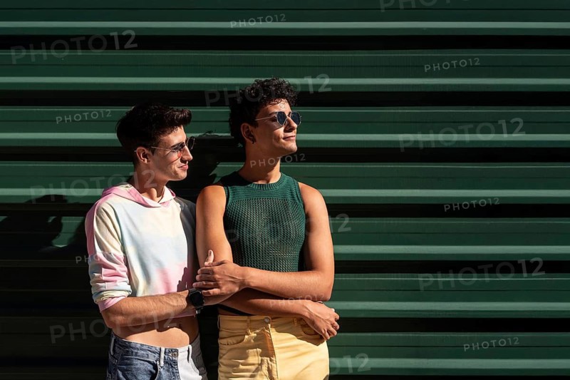 A joyful gay couple stands together, smiling in the sunlight against a green wall. They exude a sense of happiness and love, enjoying their moment in a casual setting