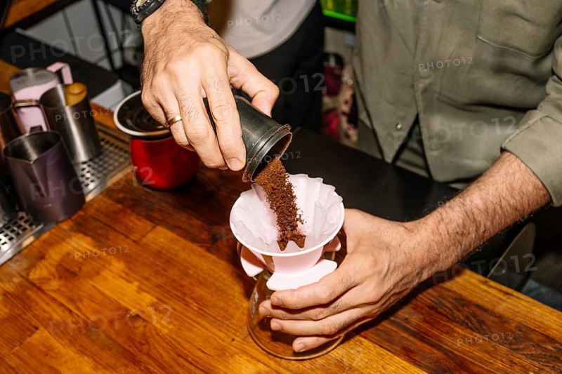 A barista pours freshly ground coffee into a paper filter placed in a pour-over coffee maker. The scene is set on a wooden counter, highlighting a specialty coffee brewing process