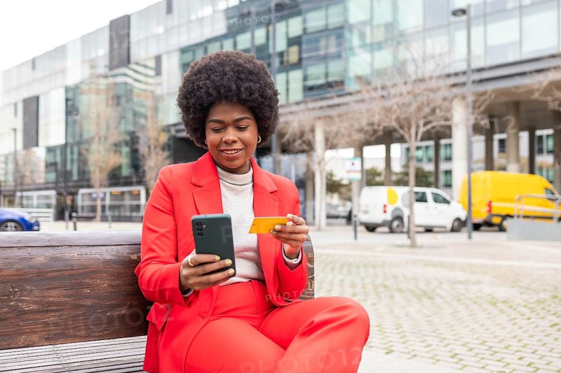 A confident woman in a red suit sits on a bench, using her smartphone and holding a credit card. The modern cityscape in the background highlights a vibrant lifestyle