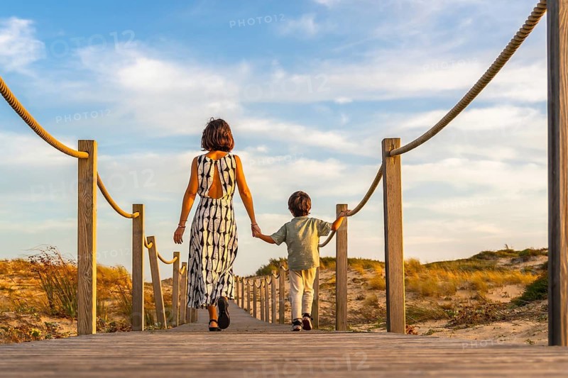 Mother and son walking hand in hand on a boardwalk through sand dunes toward the beach at sunset, enjoying family vacation moments and leisure time together