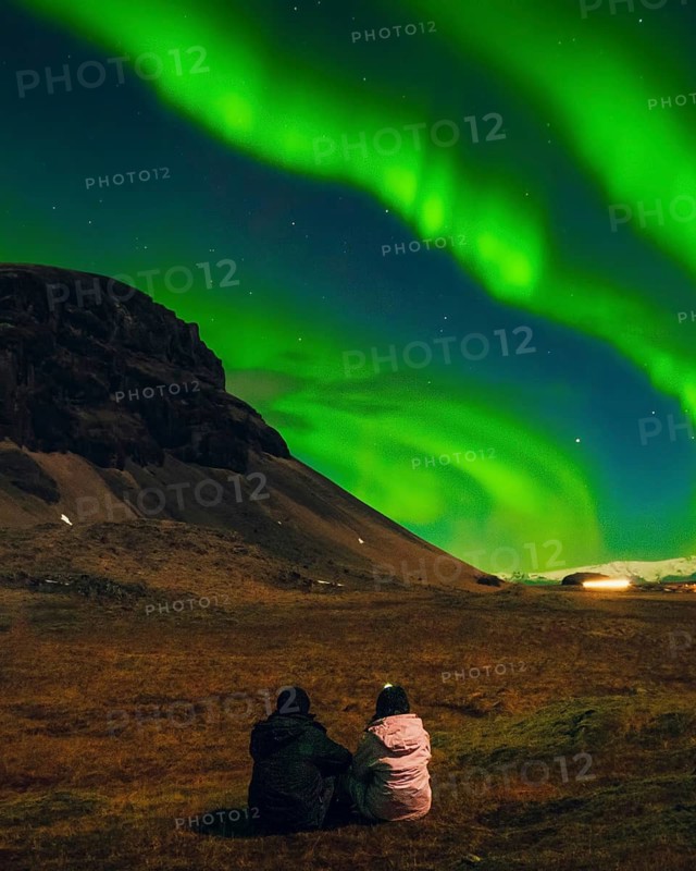 Two people romantic couple sitting under vivid aurora borealis with northern landscape, magical winter night sky with green northern lights