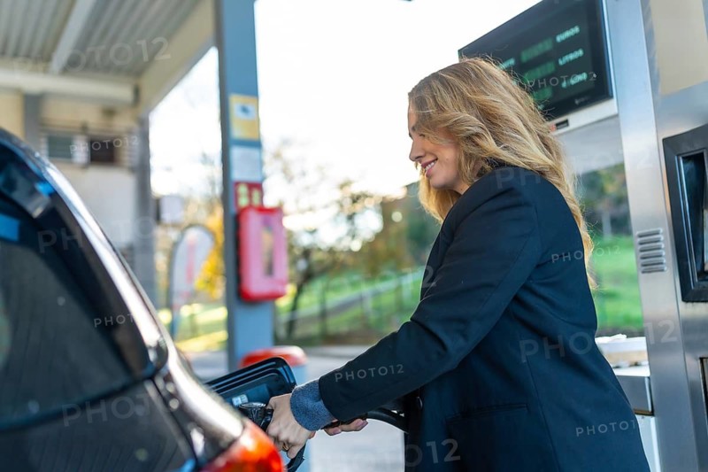 Woman refueling her car at a gas station, smiling while holding the pump nozzle everyday routine of travel, transportation and coping with rising fuel costs and expenses