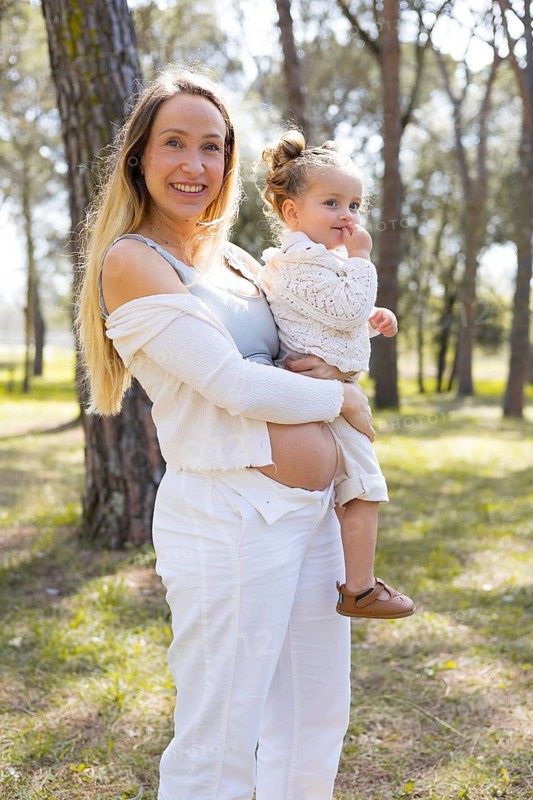 A pregnant woman stands outside in a serene natural setting, holding her young daughter. The scene captures a tender moment of family bonding and maternal love