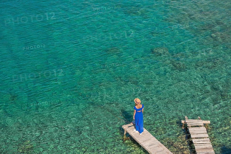 A woman in a flowing blue dress and hat stands on a wooden dock of Mandrakia, Milos island, Greece. Gazing at the clear turquoise waters below. The image captures a sense of tranquility and escape