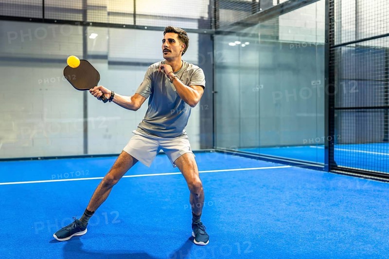 Male athlete hitting a powerful pickleball shot with a paddle on a vibrant blue indoor court, focused and dynamic, showcasing speed, agility and competitive energy